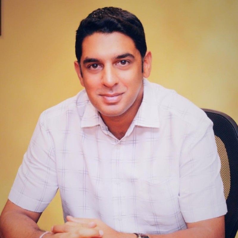 Man in a white checkered shirt smiling and sitting at a desk with a yellow background.